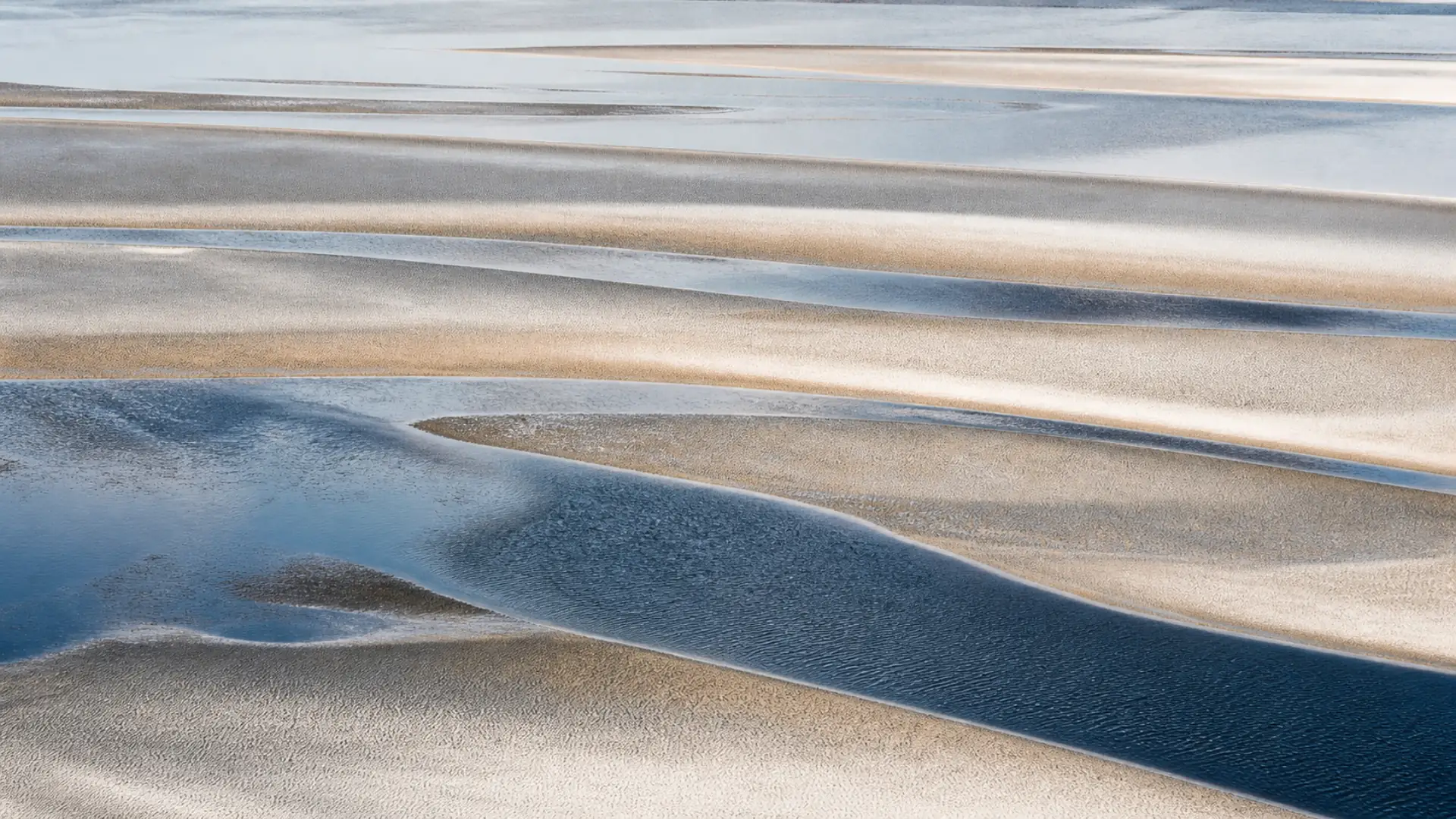 Blue tidal channels crossing pale sand in long quiet lines